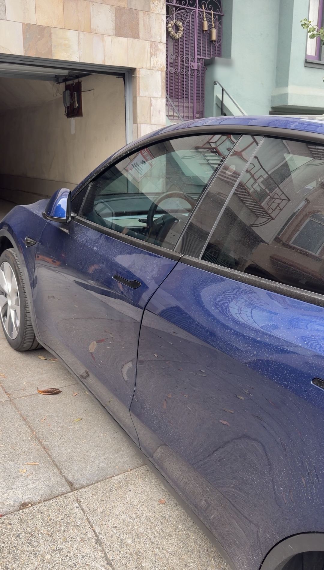 Driver's side of a deep blue Tesla Model Y before washing, paint and glass covered in dried water spots.