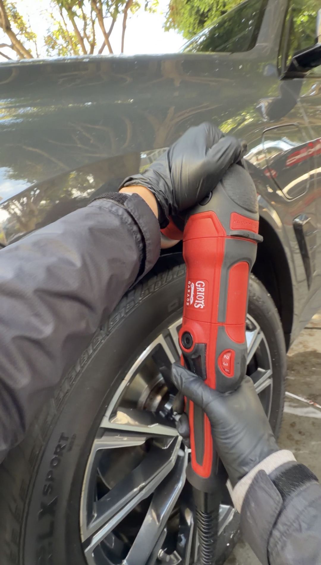 Gloved hands working a Griot's Garage polisher at the front wheel arch of a dark grey BMW X5