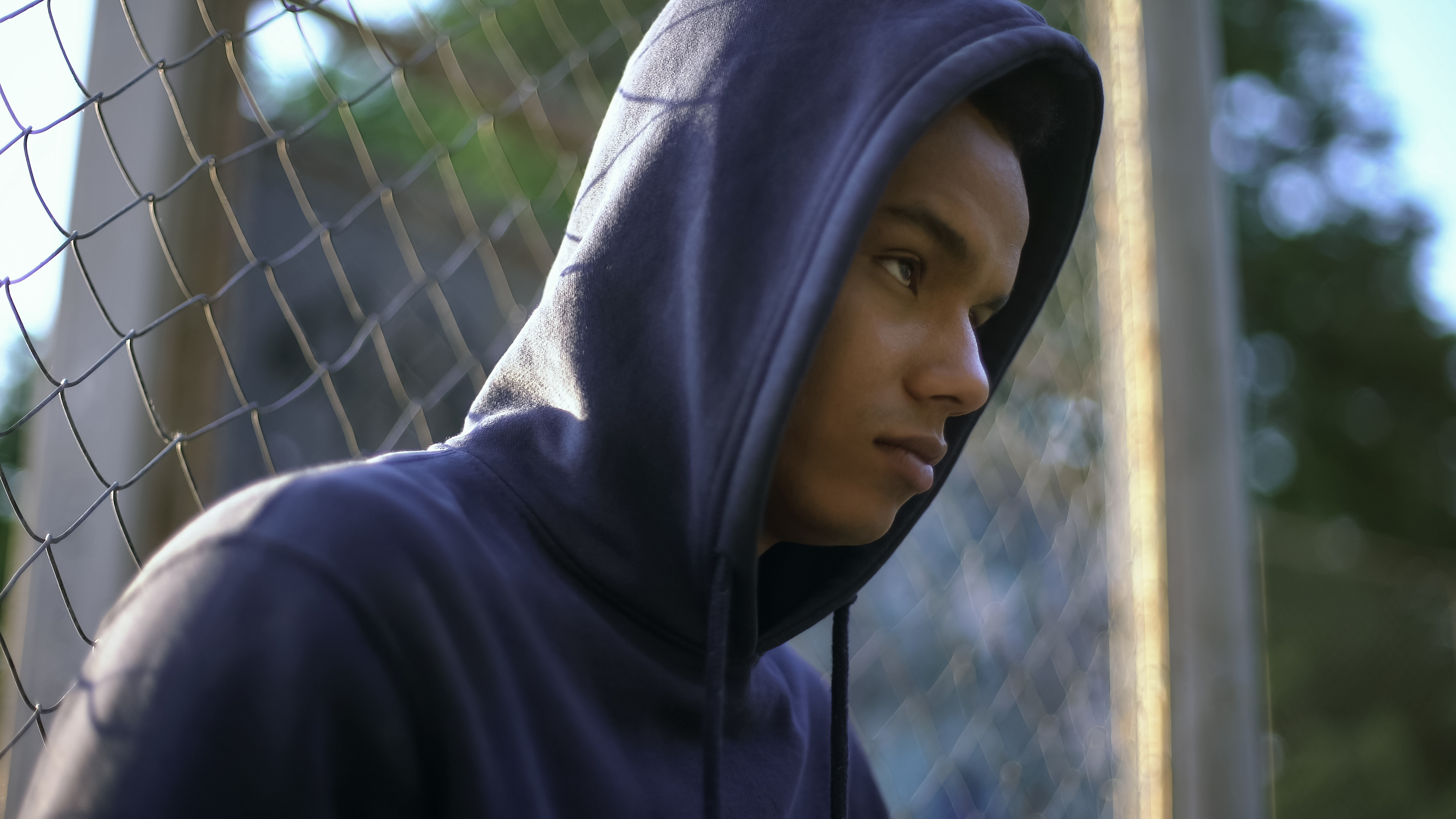 A young man in a hoodie sitting alone against a fence