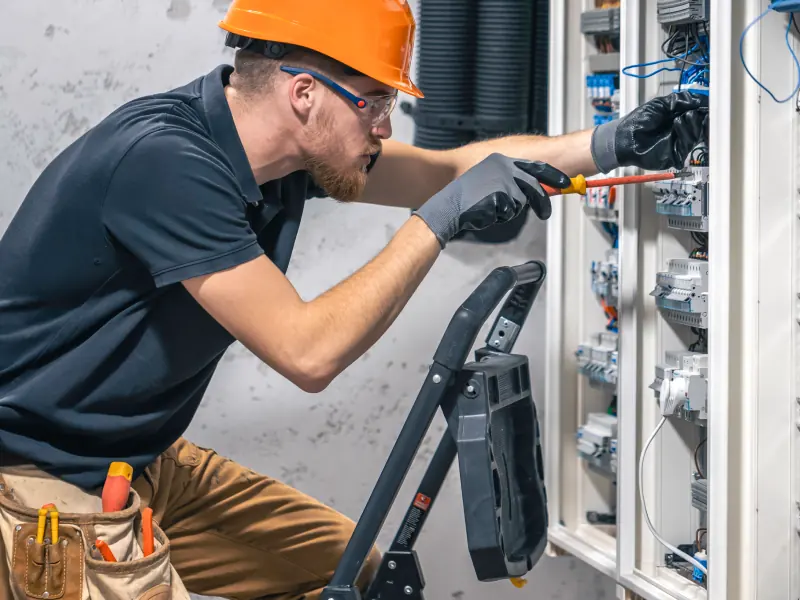 Electrician in an orange hard hat and safety glasses using a screwdriver inside a breaker panel