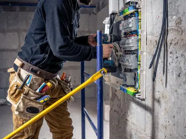 Electrician with hard hat and tool belt working on a large electrical panel in a commercial building
