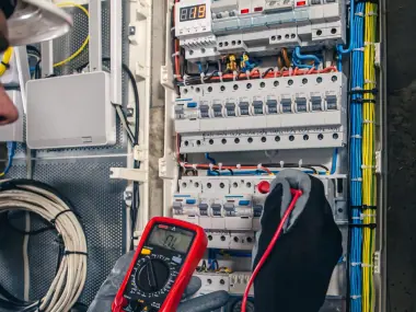 Gloved hand testing a residential breaker panel with a red digital multimeter