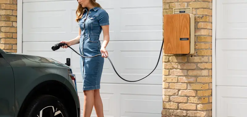 Woman plugging an electric vehicle into a wall mounted home charger beside a garage