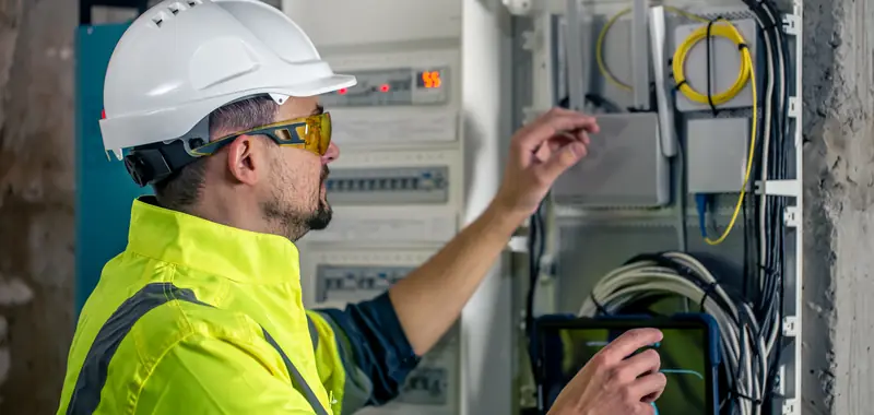 Electrician in a white hard hat and high visibility vest checking a tablet beside an electrical panel