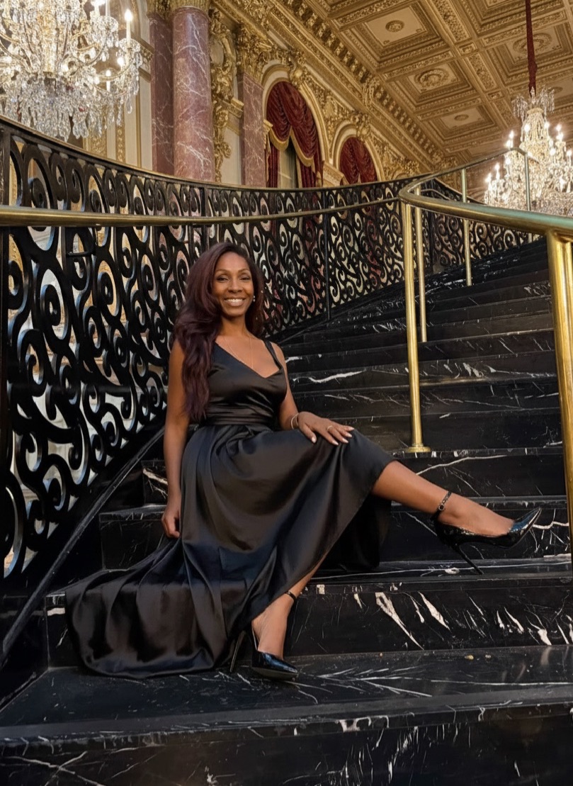 Woman wearing a long black dress sitting on a decorative staircase inside a grand building with chandeliers and ornate architectural details.