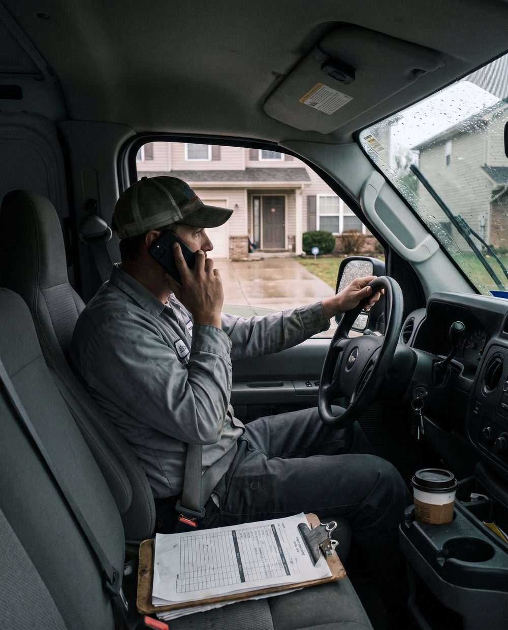An installer waiting in his van outside a house, on the phone, a job sheet on the passenger seat