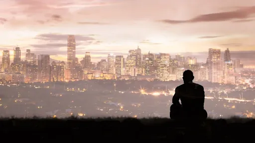 Silhouette of a person sitting alone on a hill above a city skyline at dusk