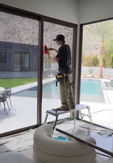 An A-Team installer squeegeeing window film onto a floor-to-ceiling sliding glass door, drop cloths over the floor and furniture.