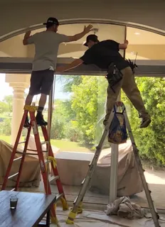 Two A-Team installers on ladders applying window film to a large arched window, with drop cloths covering the floor below.