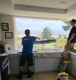Two A-Team installers fitting window film to a wide kitchen window above the sink, counters covered with drop cloths.