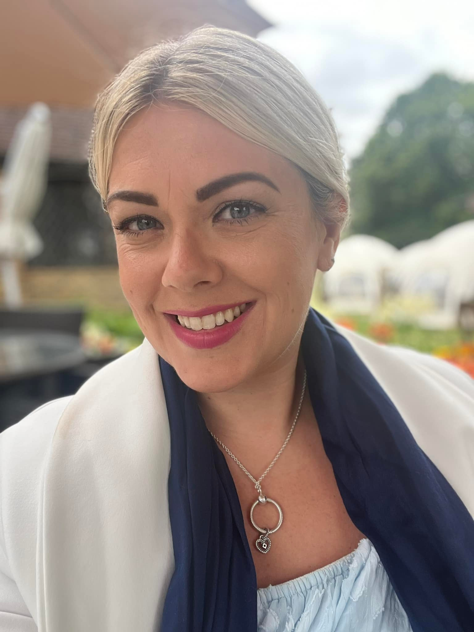 Nikki Tapley, smiling with light hair, wearing a white jacket and blue top, seated outdoors, founder of Inspired Women Mastermind.