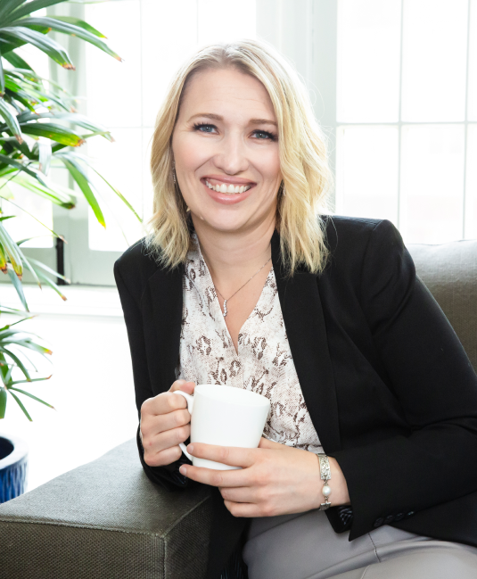 Photo of Mandi Ellefson sitting on a drk gray sofa holding a cup of coffee.