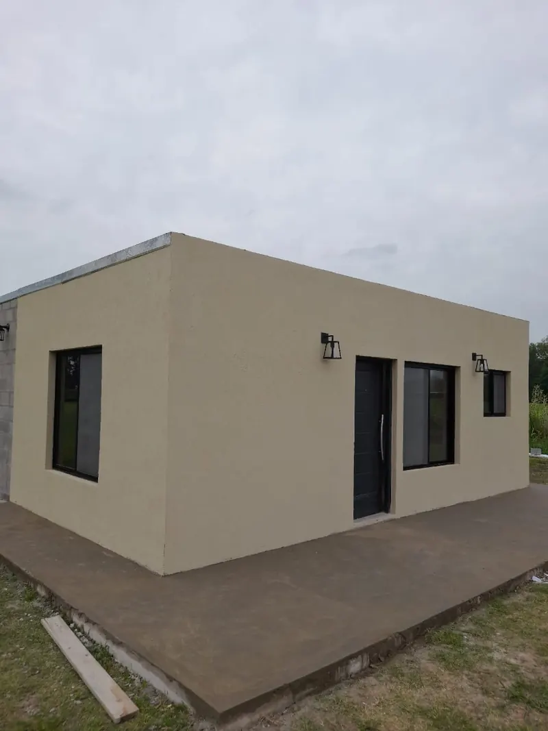 Vivienda de una planta de volumen simple, con puerta y ventanas oscuras, sobre una platea de hormigón.