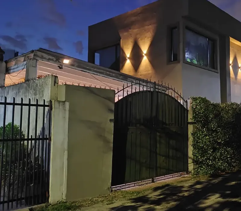 Casa de dos plantas al anochecer, con apliques de luz en la fachada y un portón negro al frente.