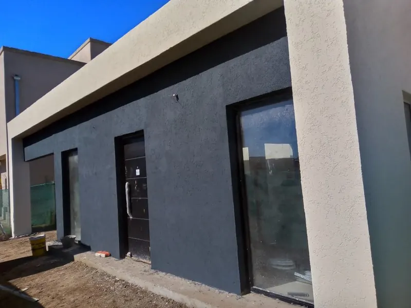 Frente de vivienda de una planta con revoque gris oscuro texturado, puerta negra y ventanal.