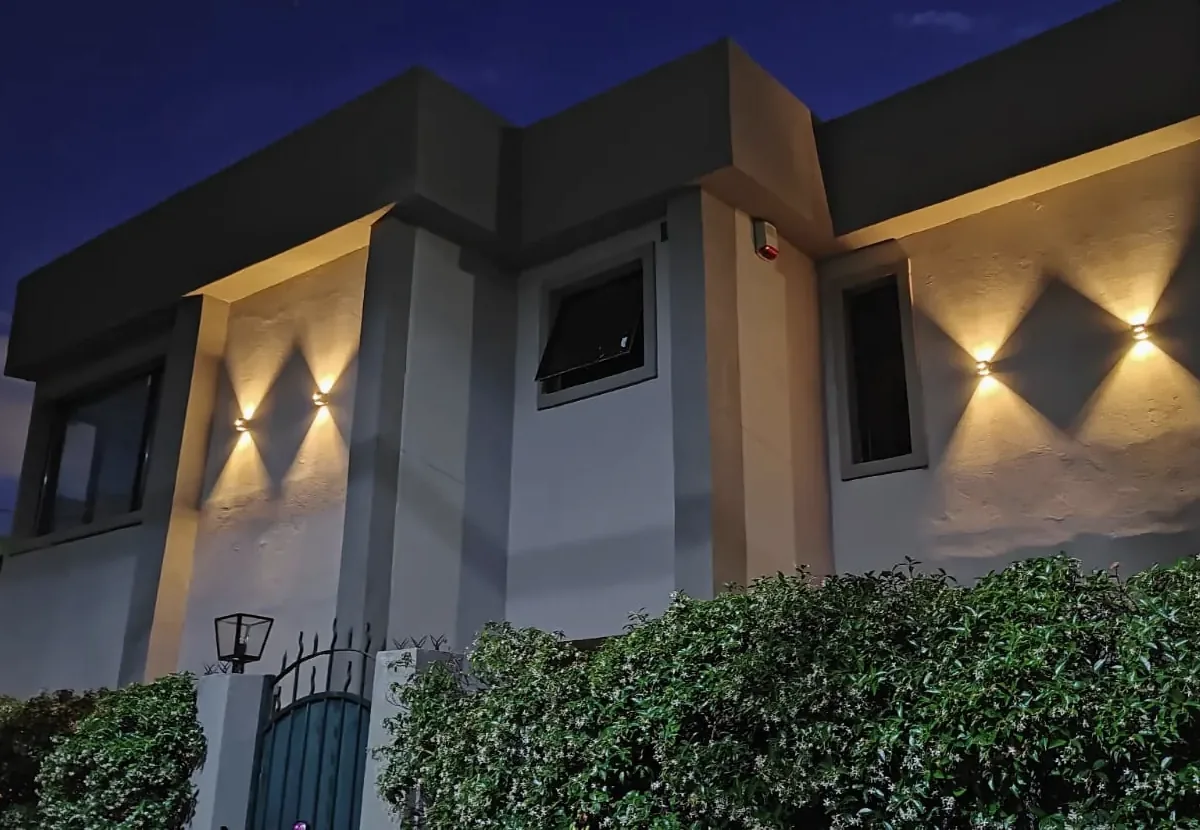 Fachada de una casa de dos plantas al anochecer, con apliques de luz que iluminan los muros claros y un cerco verde al frente.