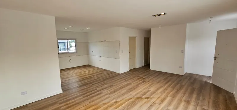 Ambiente integrado con piso símil madera, paredes blancas y espacio de cocina al fondo.