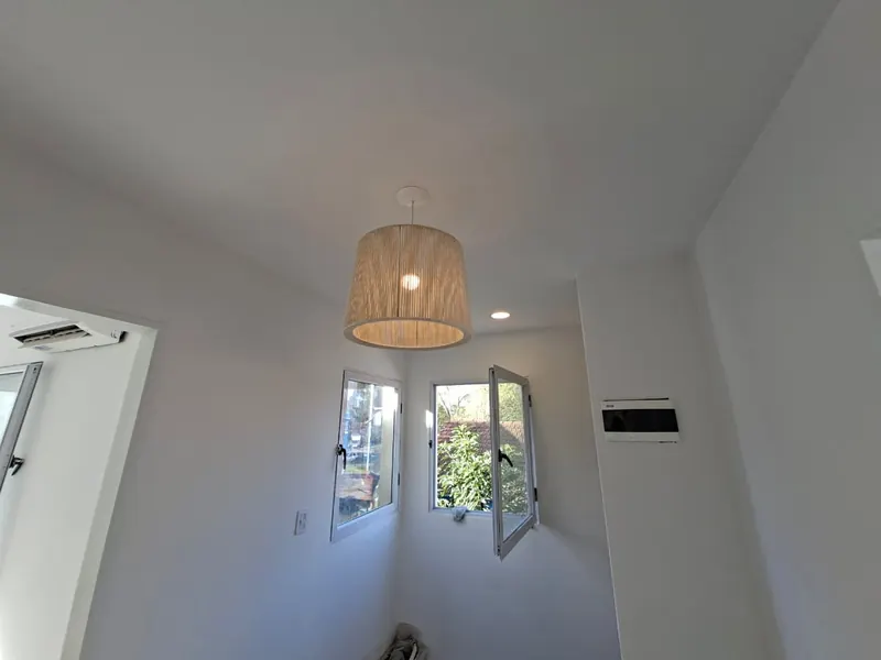 Ambiente blanco con lámpara colgante de fibra natural y dos ventanas en esquina.