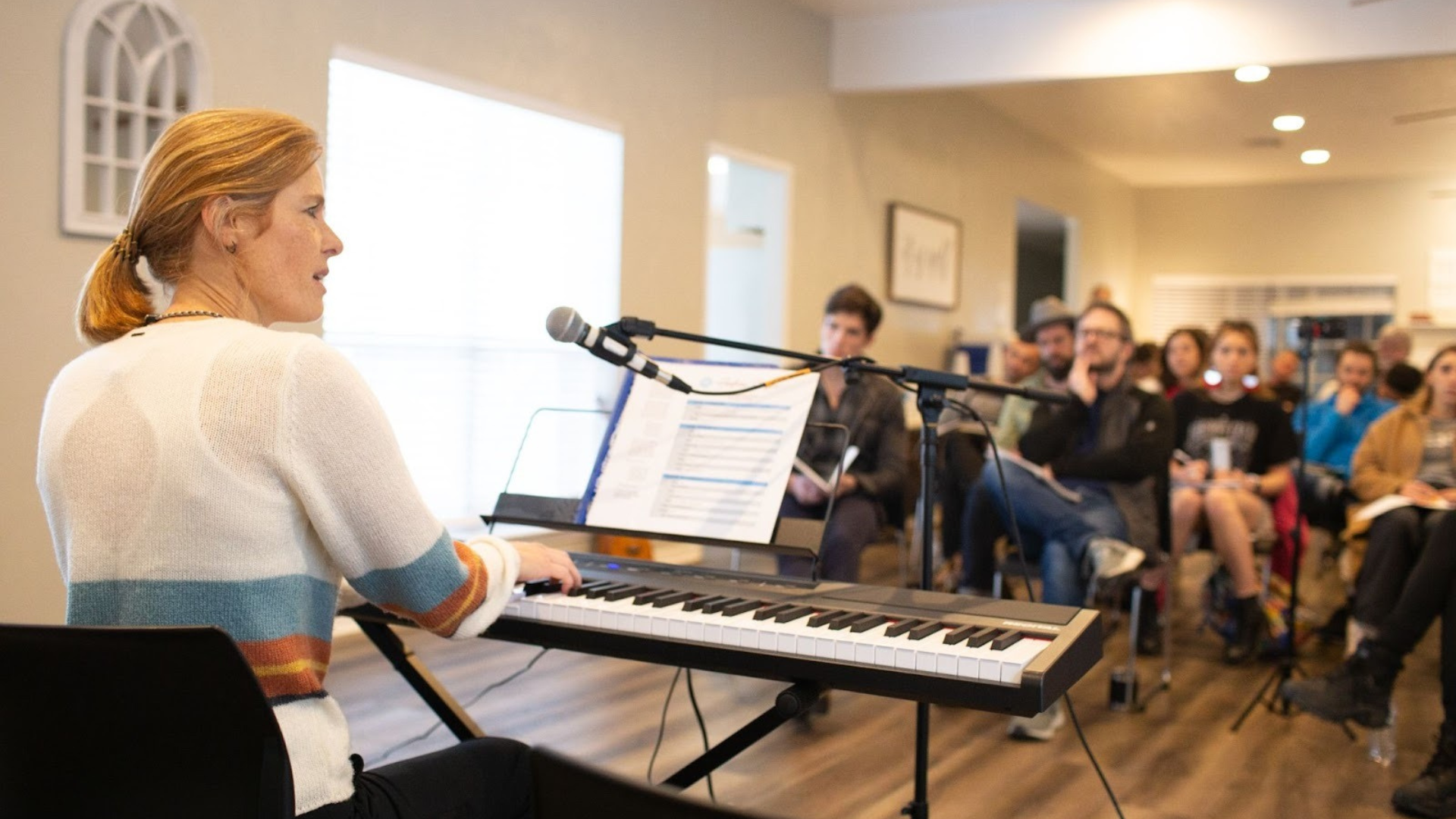 Andrea Stolpe plays a digital keyboard and sings into a microphone while leading a songwriting workshop, with an audience seated in a bright room. Andrea Stolpe plays a digital keyboard and sings into a microphone while leading a songwriting workshop, with an audience seated in a bright room.