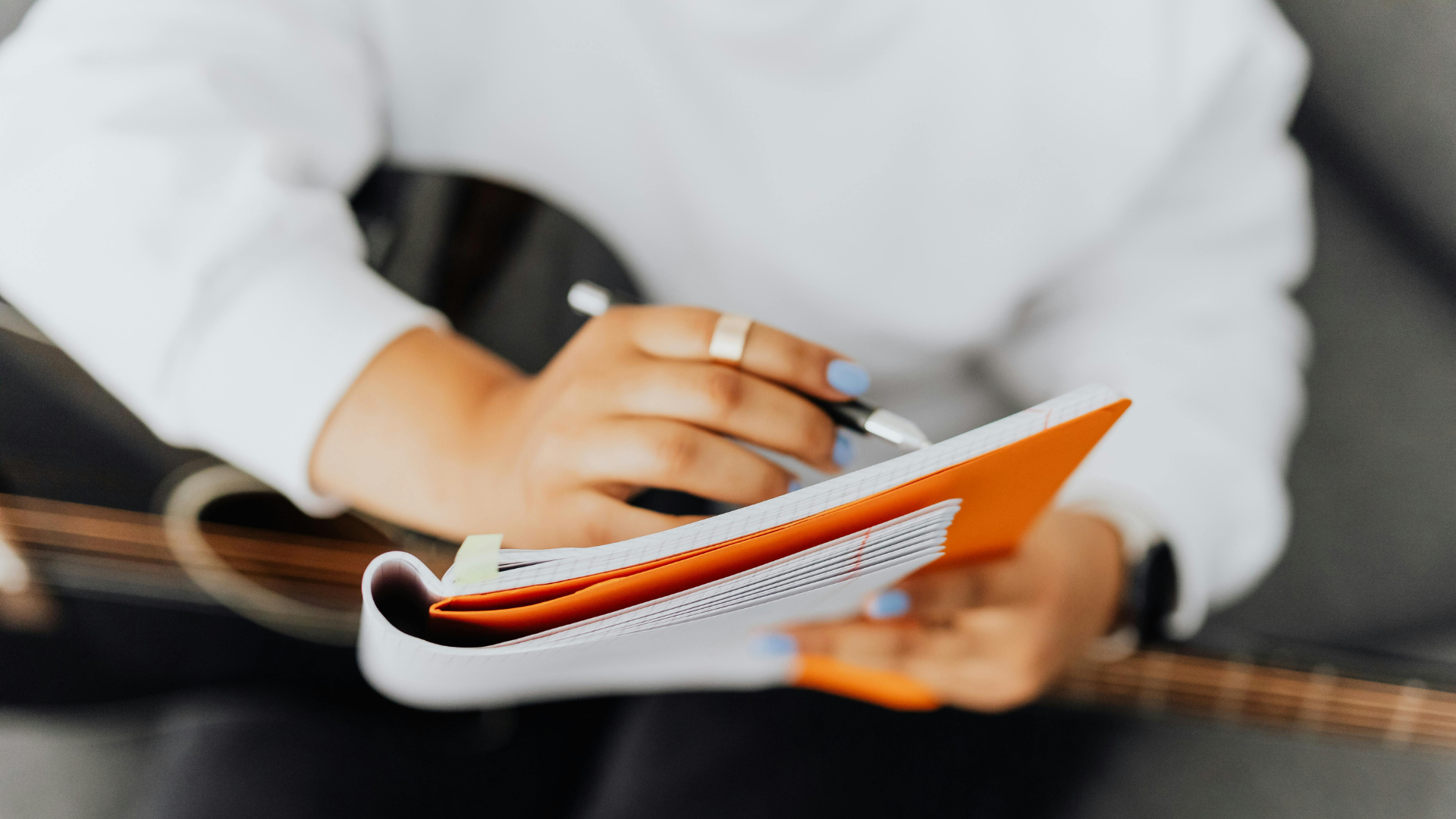 Musician's hands holding a pen and a notebook with an orange cover, focusing on the process of writing and editing song lyrics. Musician's hands holding a pen and a notebook with an orange cover, focusing on the process of writing and editing song lyrics.