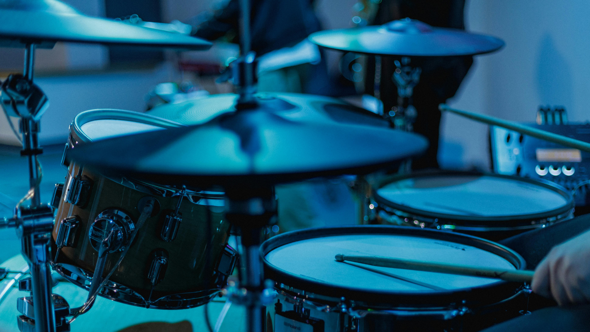 Close-up of a drum kit under blue stage lighting, with drumsticks resting on the snare and cymbals in the foreground.