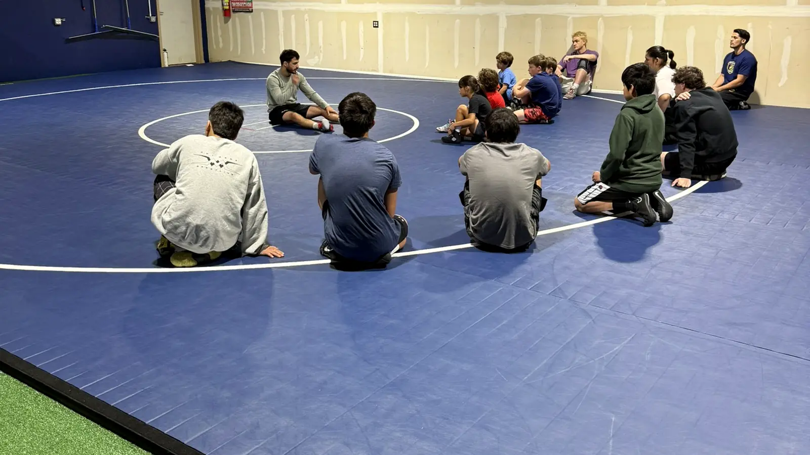 Young athletes sit in a circle on the mat listening to their coach