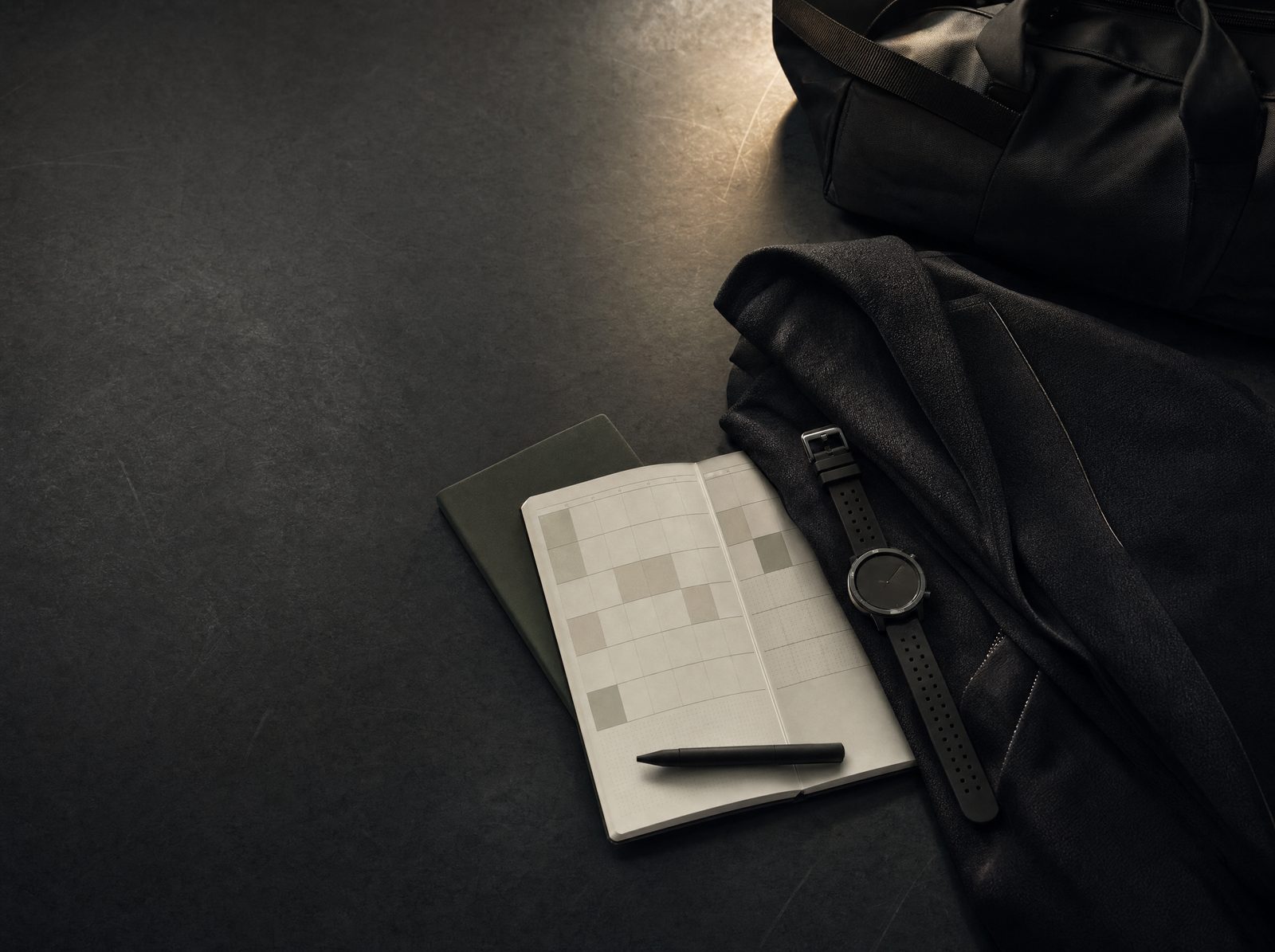 A dark editorial still life with a weekly planner, watch, tailored jacket and training bag.