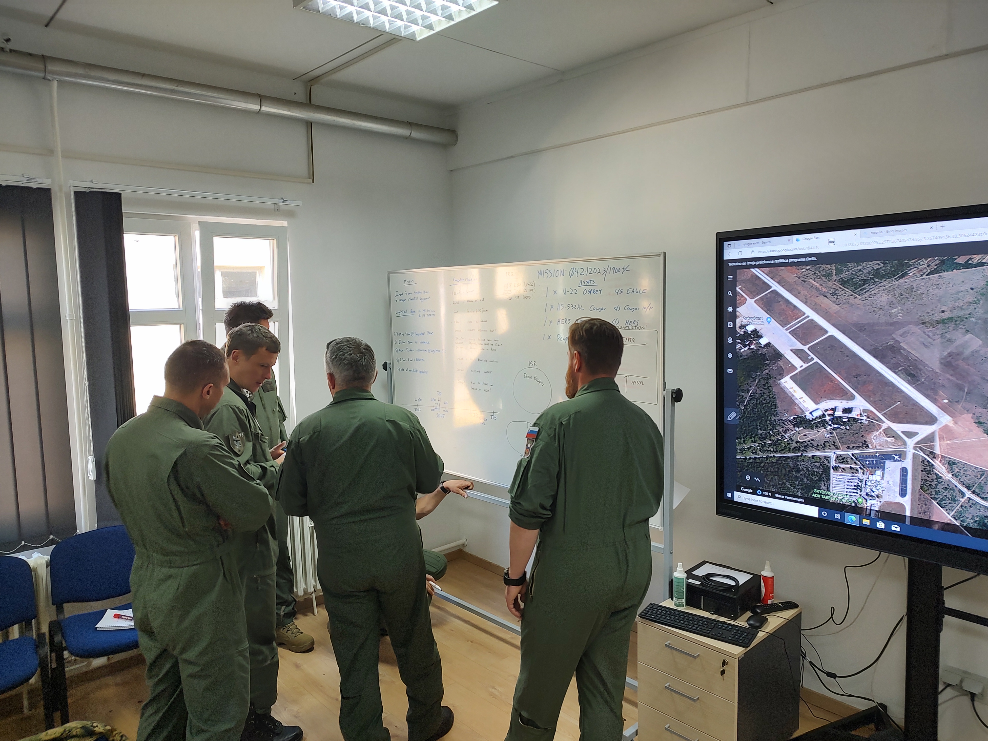 Helicopter pilots in flight suits reviewing a whiteboard briefing and airfield map during a training session
