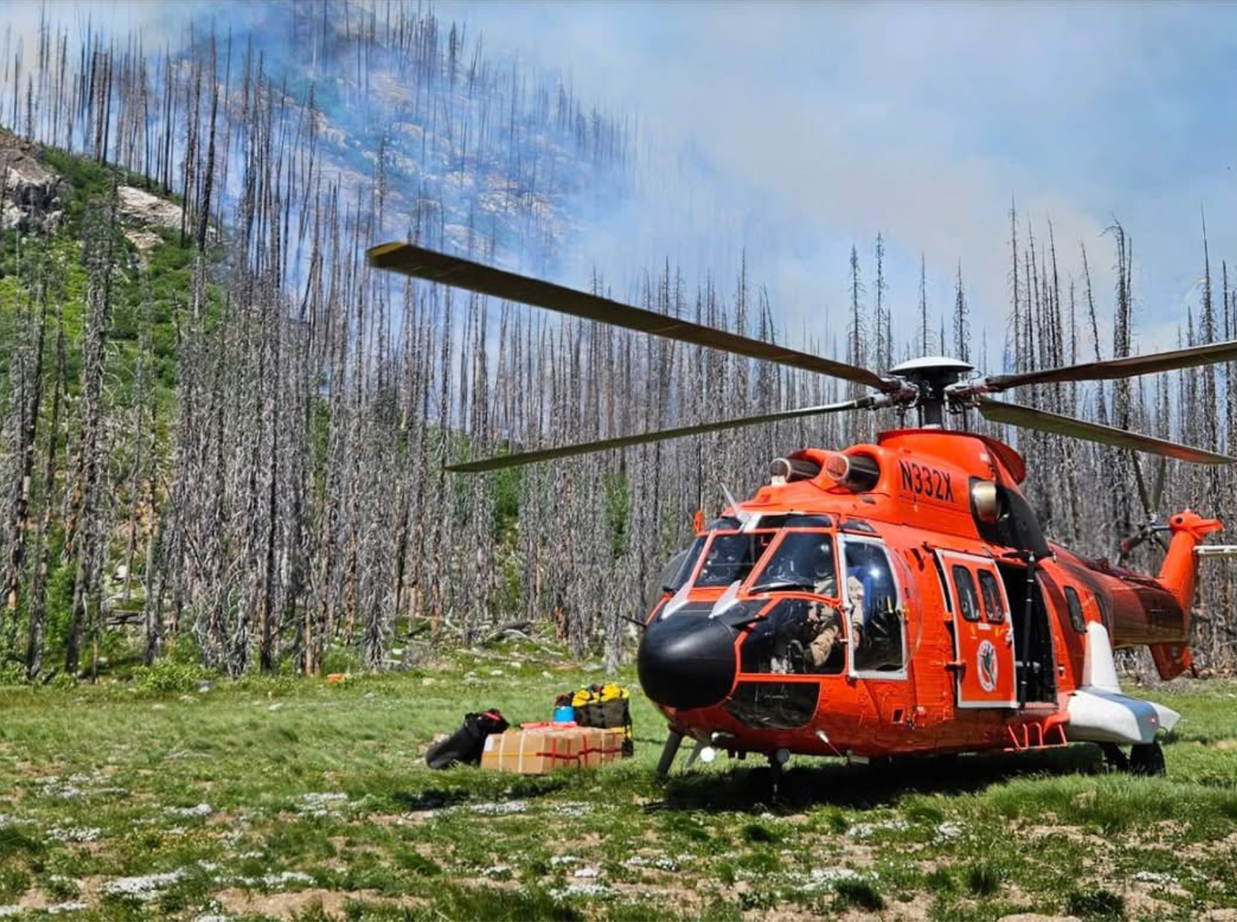 Orange helicopter landed in a clearing beside a burned forest with smoke on the ridge