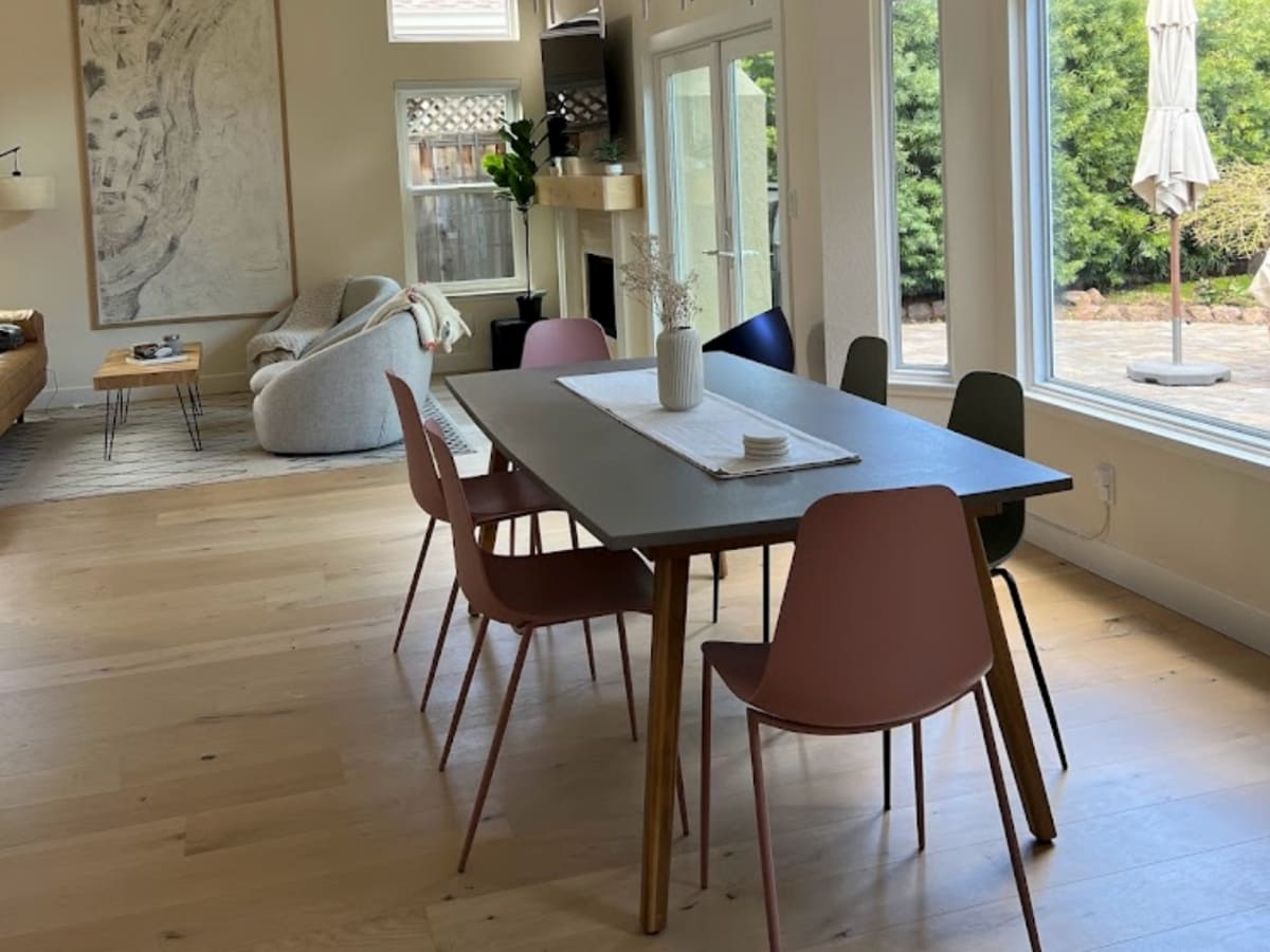 A modern dining space with a large table surrounded by colorful chairs, set in an open-plan living area with natural light pouring in from large windows.