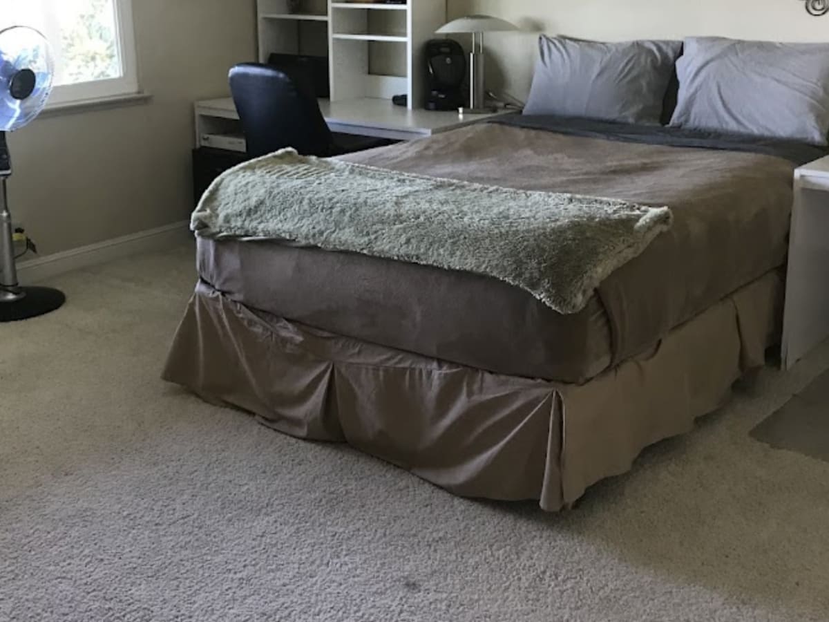 Well-maintained bedroom with a bed covered in taupe bedding and a fan by the window, representing expert carpet cleaning and bedroom organization in Napa.”