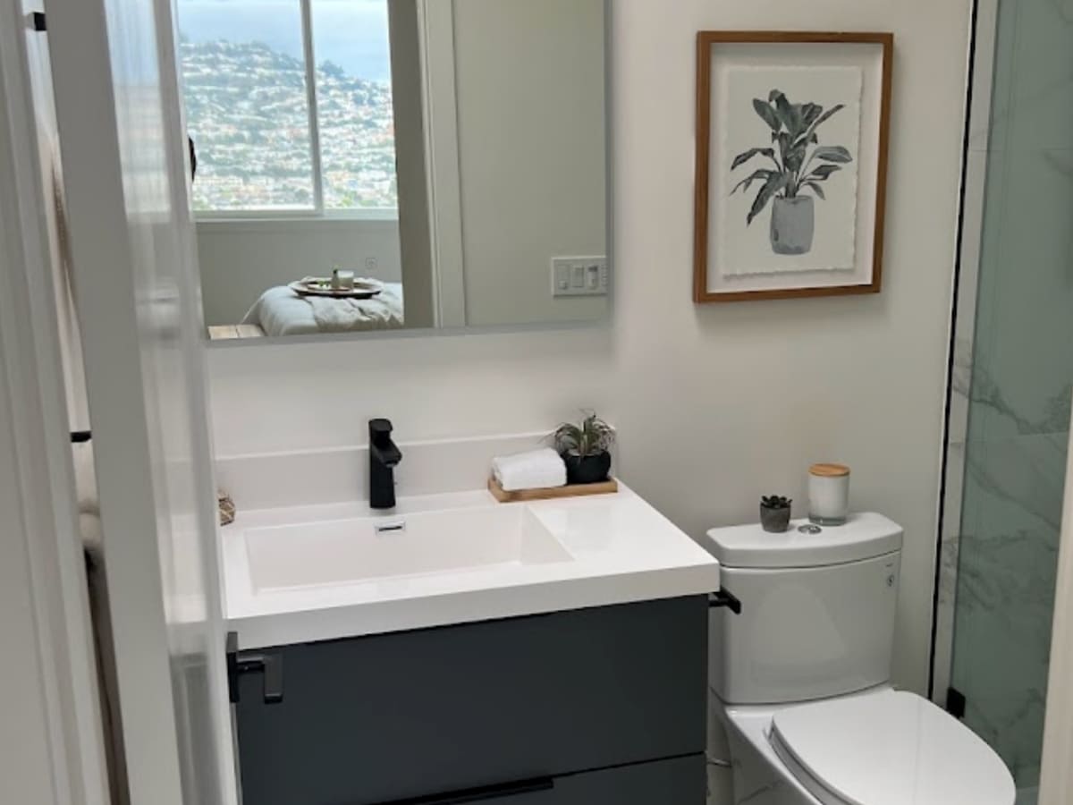 A sleek bathroom with a minimalist design, featuring a large mirror, a small plant on the counter, and a view of the city through a window in the adjacent bedroom.