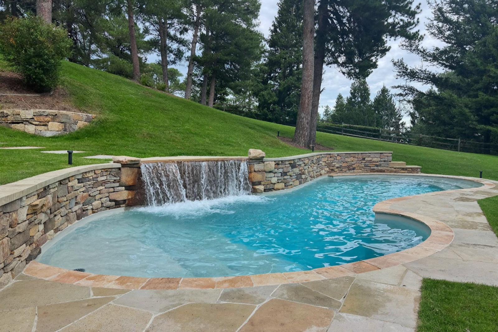 Freeform pool built into a sloped yard with a stacked stone retaining wall and waterfall