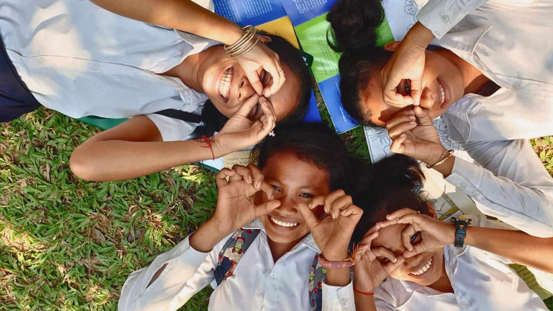 A group of young girls lying on the grass, each making the sign of the cross with their hands