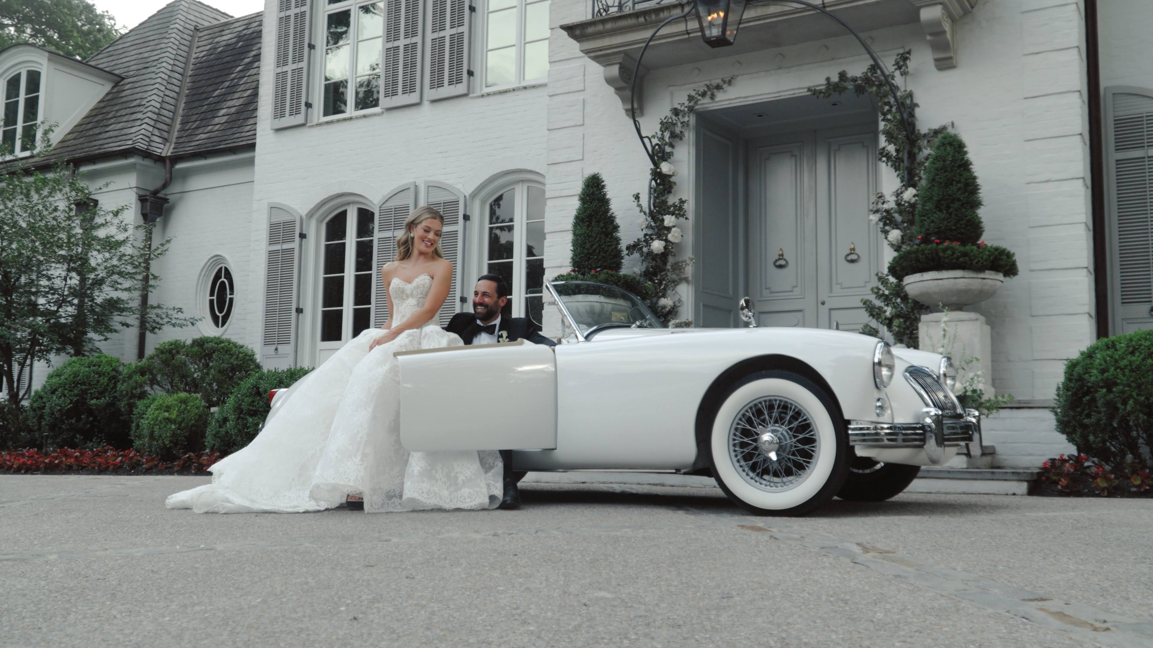 A newlywed couple in the Getaway Car, a restored white vintage MGA roadster, outside a grand estate