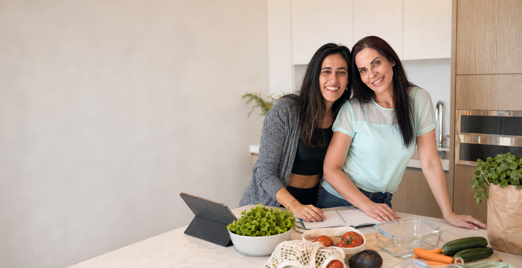 Victoria and her mother Mirna standing together at a bright kitchen island with a tablet, an open planner and fresh produce.