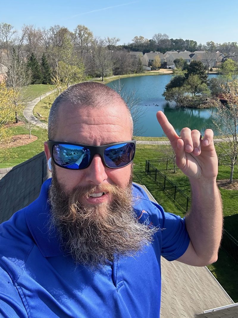 John wearing sunglasses on a roof, pointing up, with a pond and neighborhood behind him