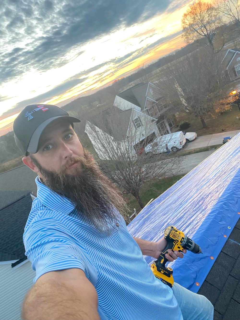 John, the Bearded Roofer, on a roof holding a drill next to a blue tarp at sunset