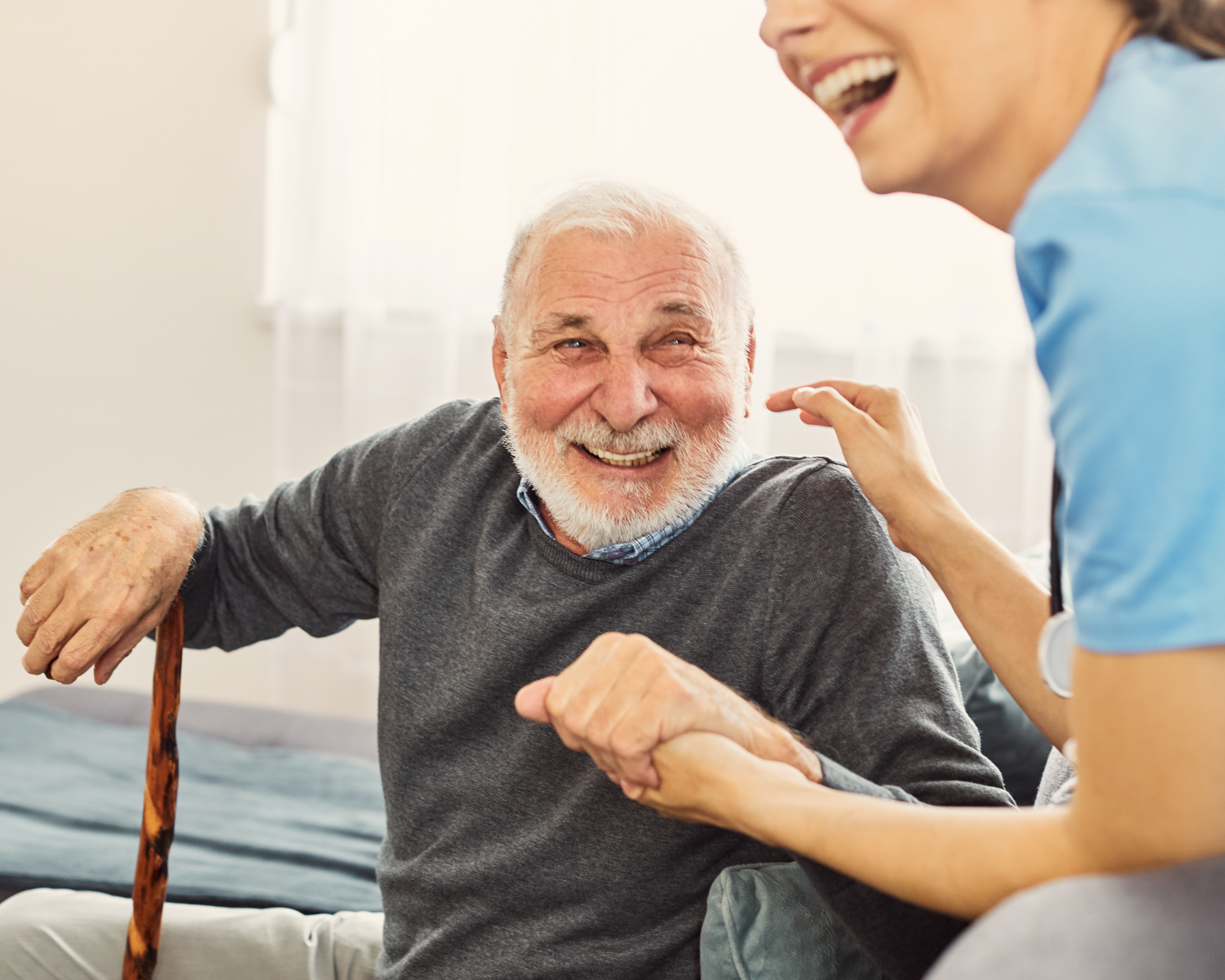 Adult caretaker nurse helping a senior man while laughing in a warm, sunlit room
