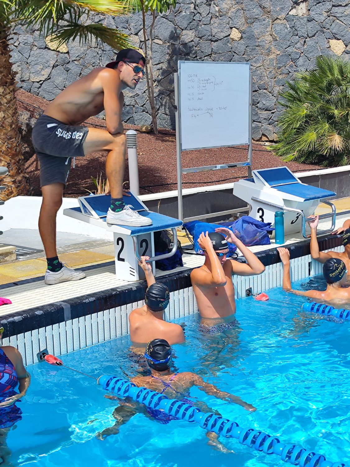 Swimpros coach talking a junior group through the set from the starting block, whiteboard by the pool in Tenerife