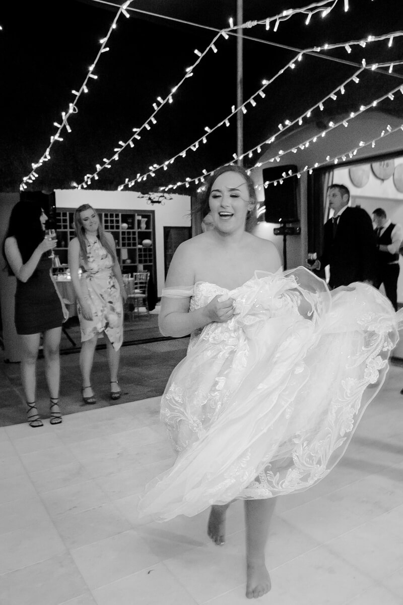 A barefoot bride laughing as she lifts her dress on the dance floor under fairy lights