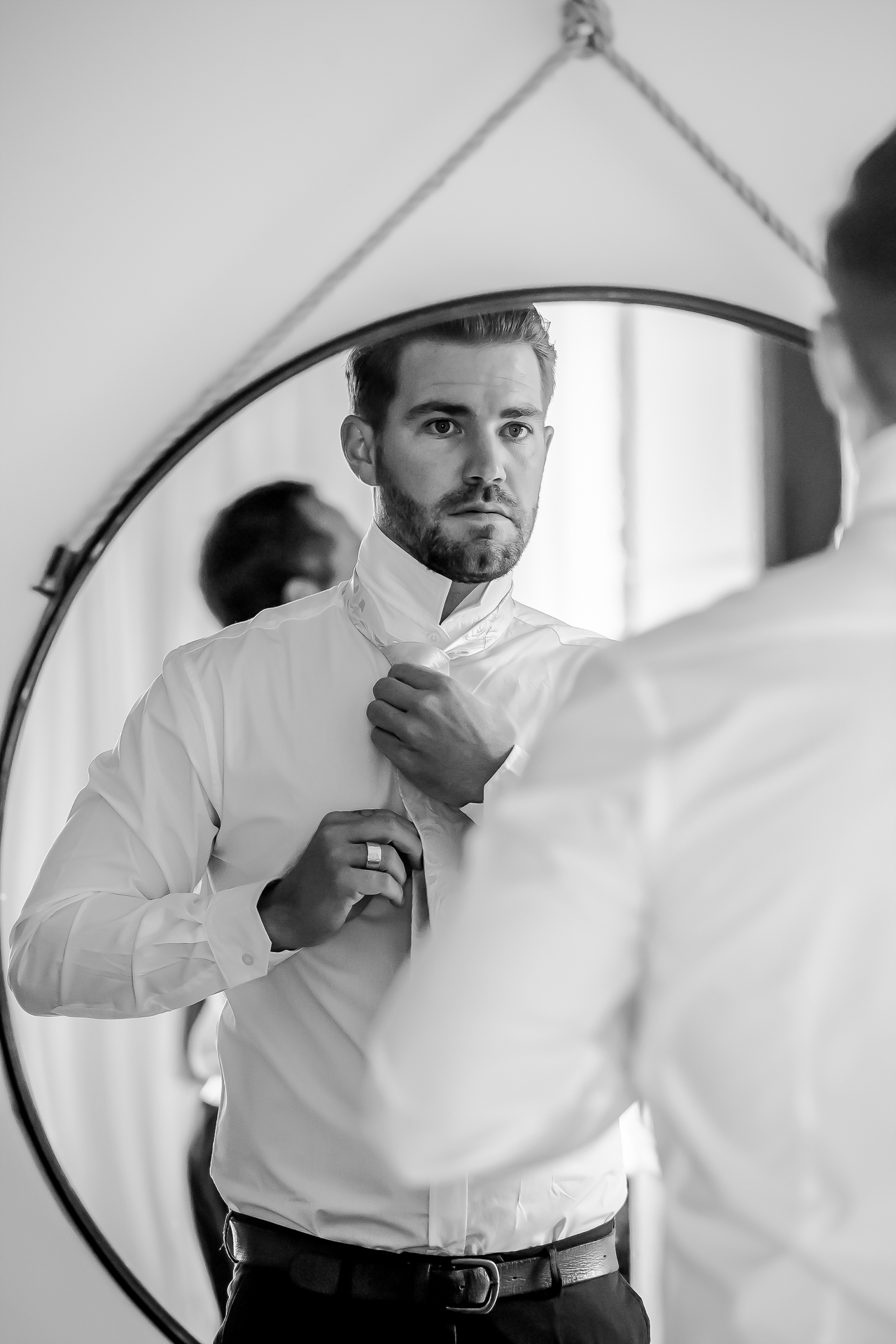 A groom doing up his tie in a round mirror hanging from a rope