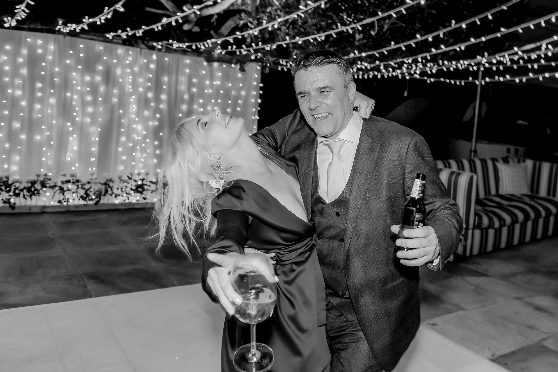 A man with a beer dancing with a guest under fairy lights