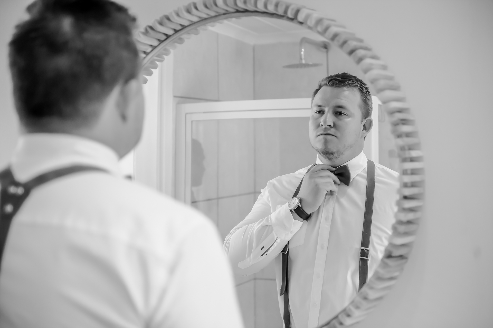 A groom fixing his bow tie in a round mirror