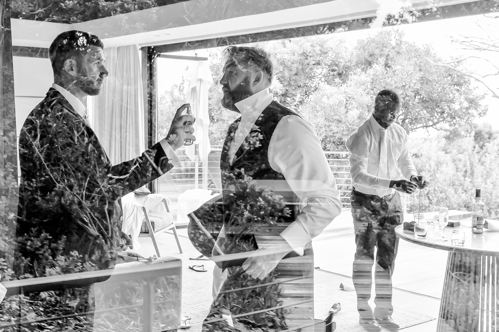 A groomsman spraying cologne on the groom, seen through glass