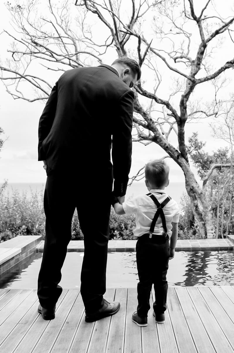 A man in a suit holding a little page boy's hand on a deck above the sea