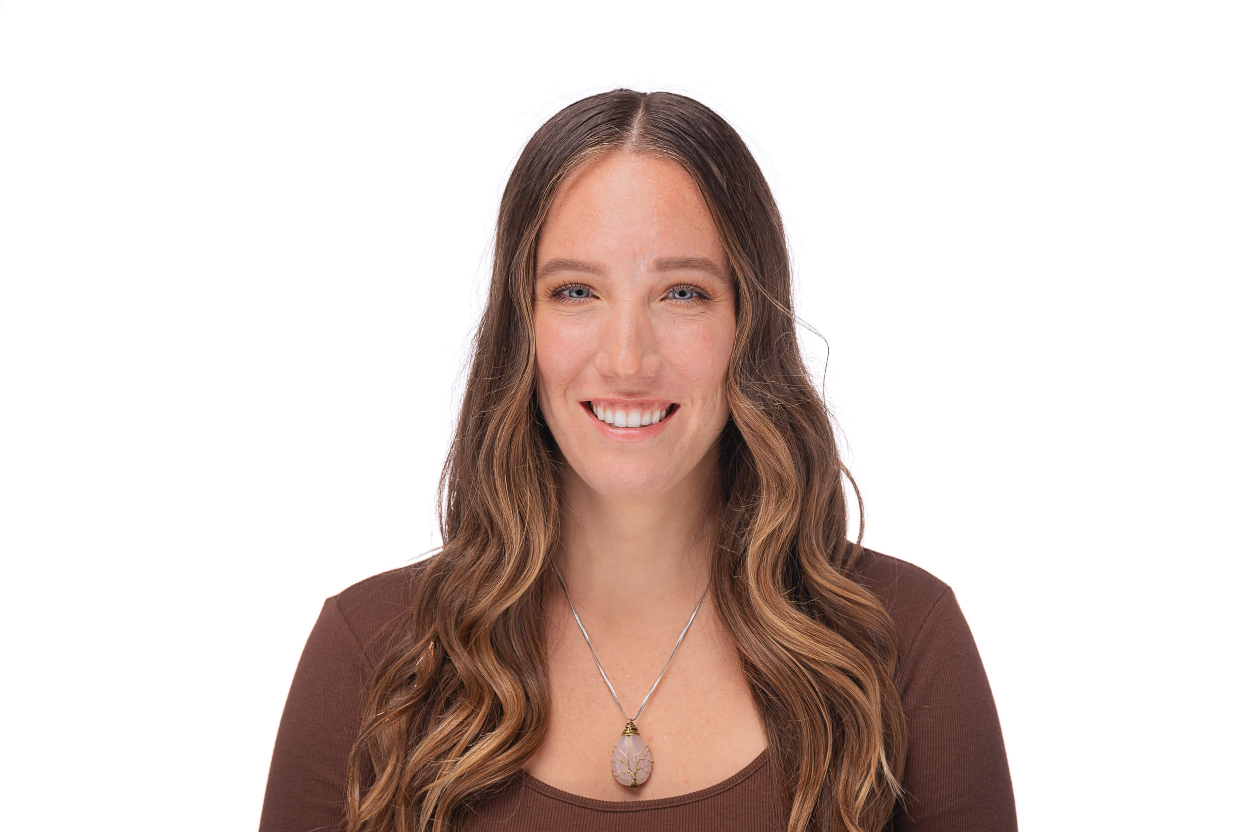 A professional headshot featuring a woman with long, wavy chestnut hair, wearing a simple brown top and a pendant necklace. She is smiling gently and looking directly at the camera with a friendly expression, set against a clean white background.