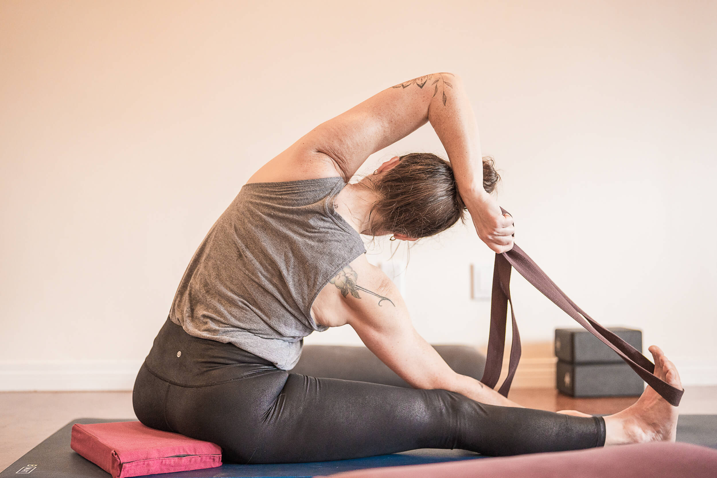 A serene and focused individual practicing a yoga pose in a Shala Yoga studio. The yogi, with visible tattoos, is using a strap to deepen a seated forward bend, showcasing flexibility and poise. The tranquil and minimalistic studio space in Squamish provides an intimate and peaceful environment for personal growth and practice.