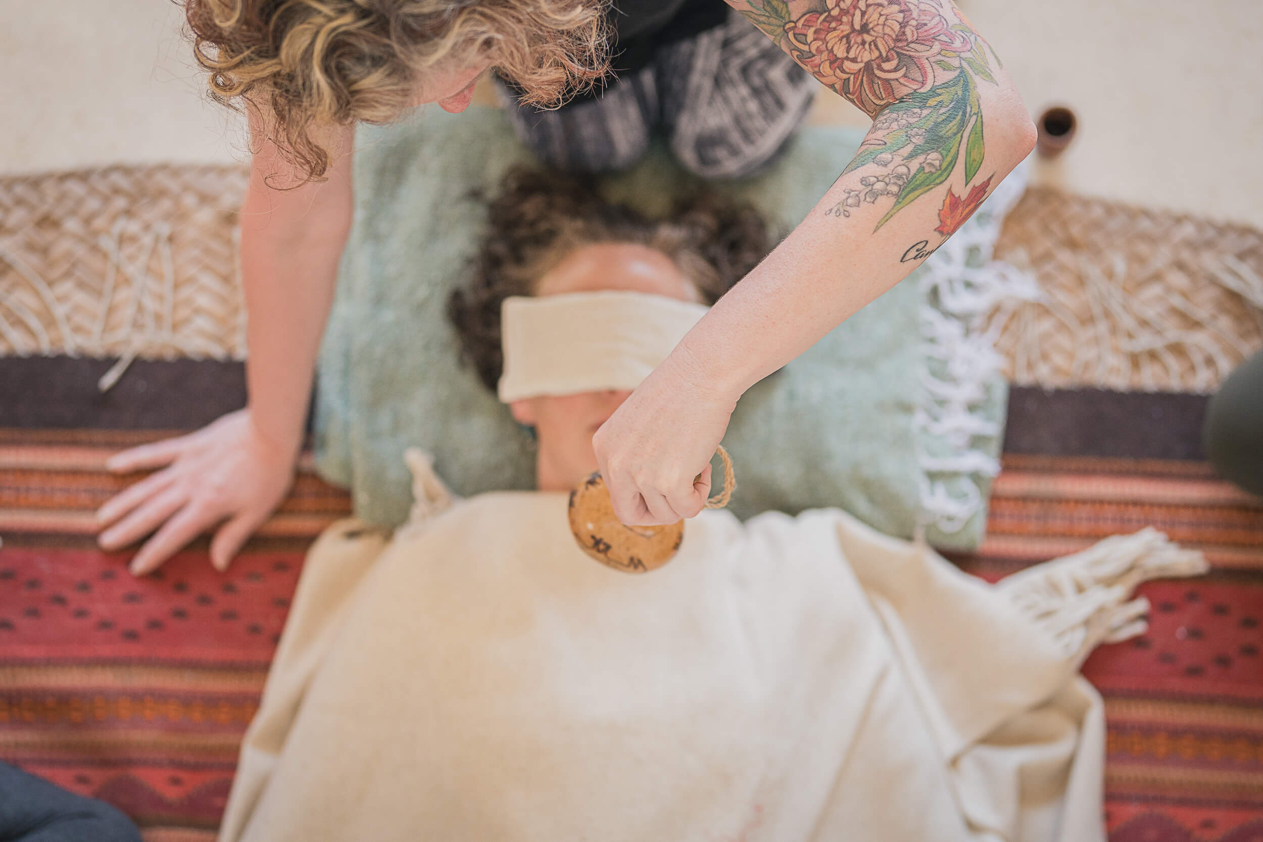 Lifestyle photographer captures a top-down view of a sound healing session with a woman receiving therapy through a singing bowl, highlighting the therapist's tattooed arm.