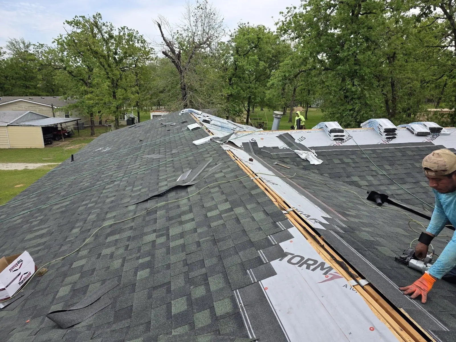 Roofer installing new shingles beside the ridge vents on a home surrounded by oak trees